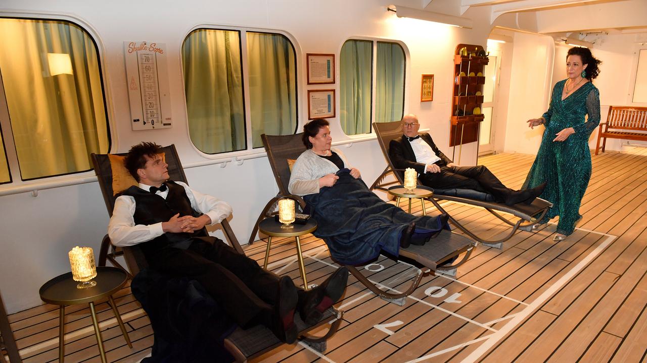 Peter Keller, Emilia Gronewald und Oliver Stiller liegen abends in Liegestühlen auf einem Schiffsdeck. Hanna Liebhold im Abendkleid steht besorgt daneben.