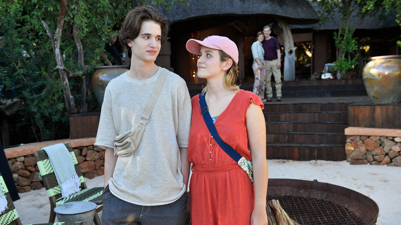  Lisa Sommer und Paul Wagner stehen im Außenbereich einer Lodge mit Sitzgelegenheiten im Sand, im Hintergrund sind ihre Eltern zu sehen.
