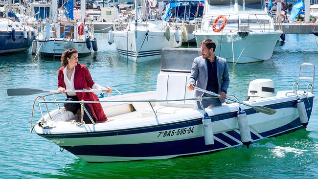 Fina und Xavi auf einem kleinen Motorboot im Hafen, umgeben von Segelbooten und Yachten, beide halten jeweils ein Paddel in den Händen