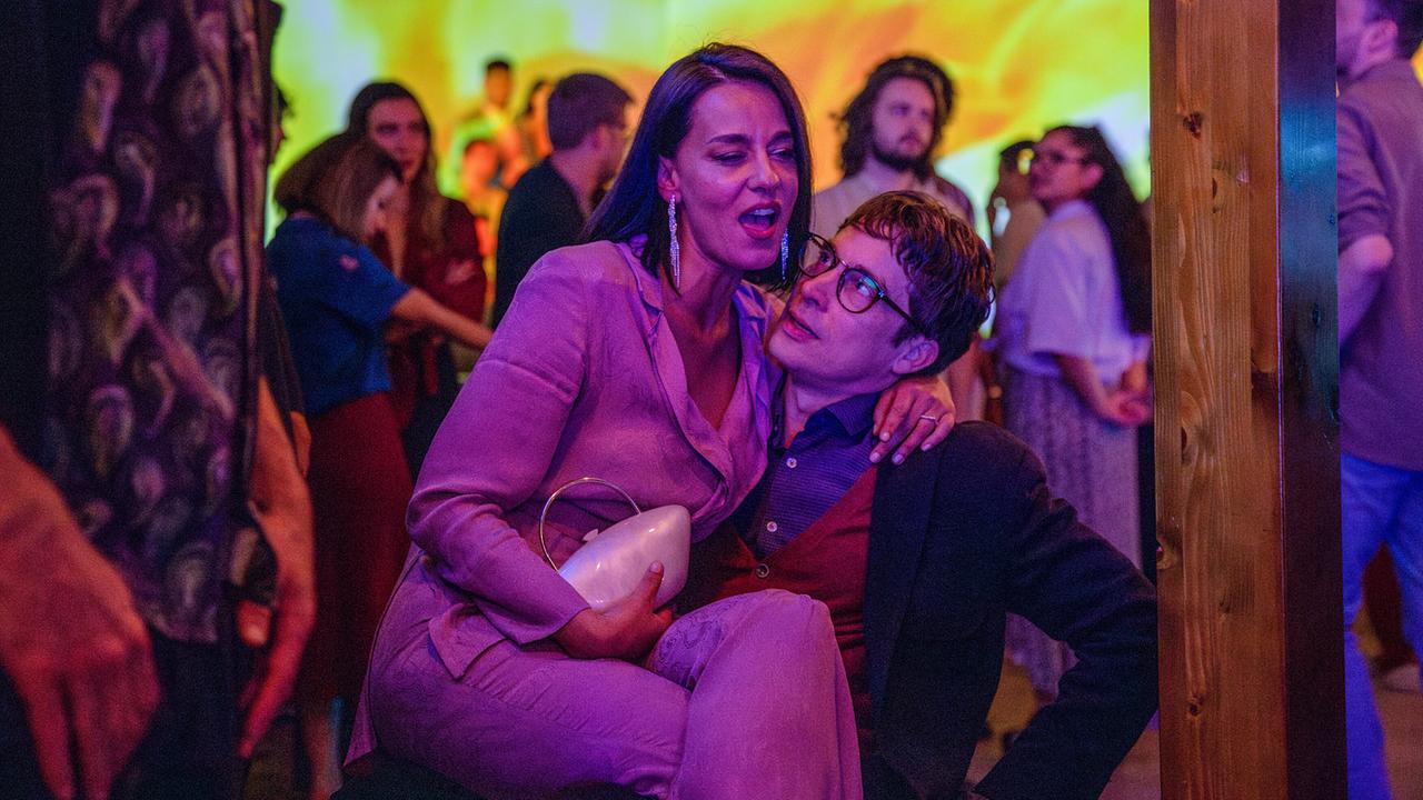 © ORF/Beta Film/Degeto/Victor Bello Bei einer Party sitzt Edita Malovčić als Sofia im eleganten Hosenanzug auf dem Schoß von Nico Rogner als Sergi mit Anzug, Hemd, Weste und Brille