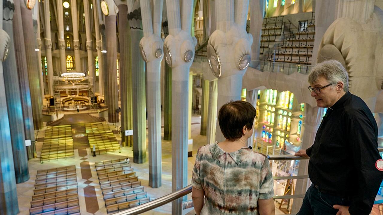 Konstantin Reymaier im Gespräch mit Rosa Ma Alsina-Pagès im lichtdurchfluteten Innenraum der Sagrada Família mit hohen, säulenartigen Pfeilern, bunten Glasfenstern und Blick auf den Altarbereich von einer Empore aus.