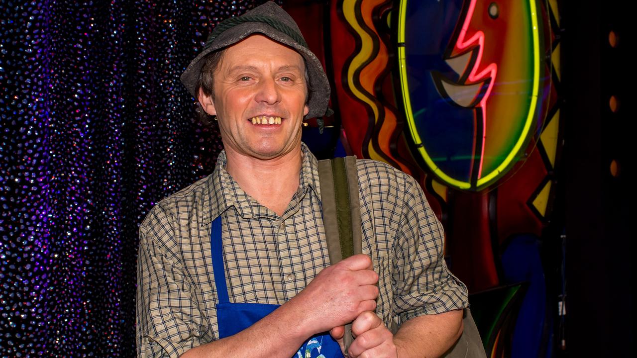 "Navi, Luis aus Südtirol" Manfred Zöschg in kariertem Hemd und blauer Schürze steht vor einer bunt beleuchteten, künstlerisch gestalteten Bühnenkulisse und hält einen Rucksackriemen fest.