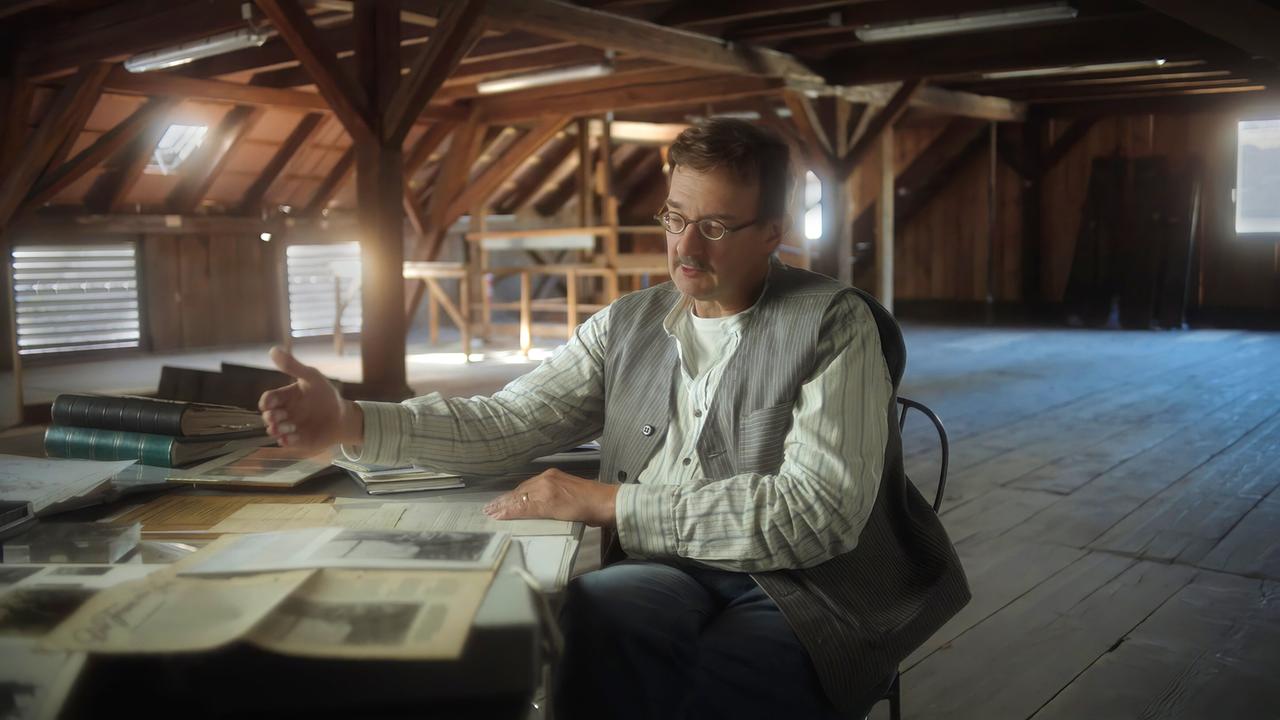 Mann mit Schnauzbart und Brille sitzt auf Stuhl und spricht über Archivakten vor ihm auf dem Tisch