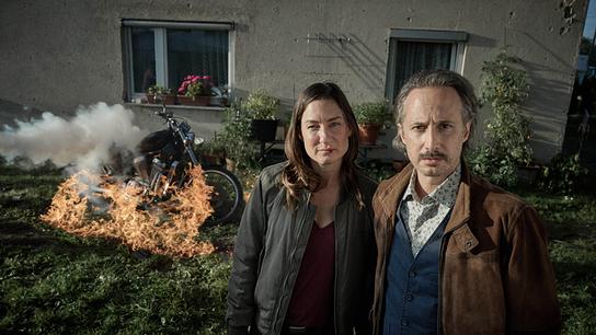 Frederike Bader und Ferdinand Zankl stehen abwartend vor einem brennenden Motorrad. 