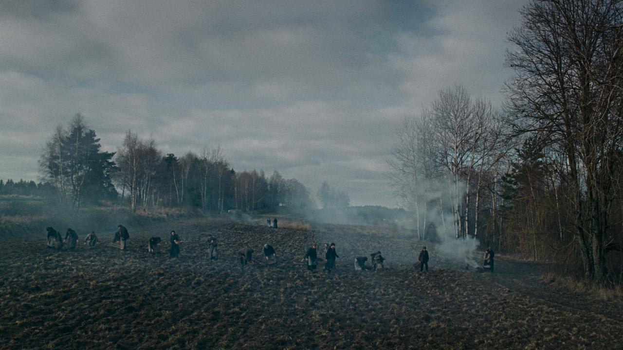 © ORF/Ulrich Seidl Film Mehrere Frauen arbeiten in einem kargen, großen Feld mit Rauchstellen unter grauem Himmel.