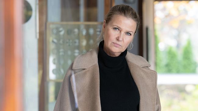 Vera Lanz in einem langen beigefarbenen Mantel steht in einem Innenraum und blickt nach vorne.