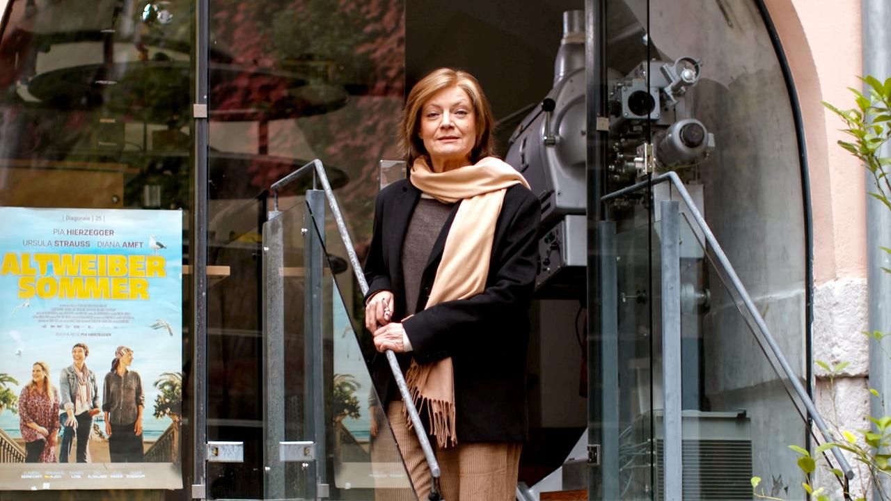 Schauspielerin Marion Mitterhammer steht auf einer Treppe vor dem Eingang eines Kinos mit Glasfassade neben einem Filmplakat zu "Altweibersommer".