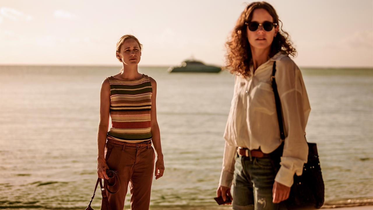 Zwei Frauen stehen in sommerlicher Freizeitkleidung am Strand, hinter ihnen ist das Meer und unscharf ein Motorboot zu sehen