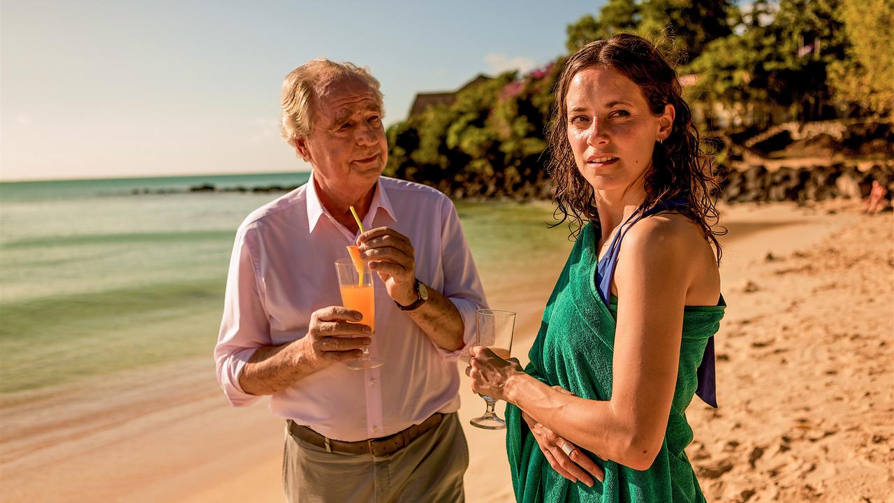 Doktor Mantler und Anaja Knauer stehen am Strand und halten Cocktail-Gläser in den Händen.