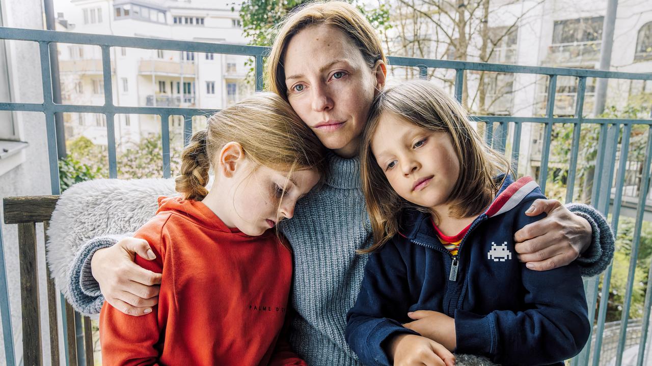 Ines hält ihren beiden Kindern Emma und Sammy im Arm auf einem Balkon in städtischer Umgebung