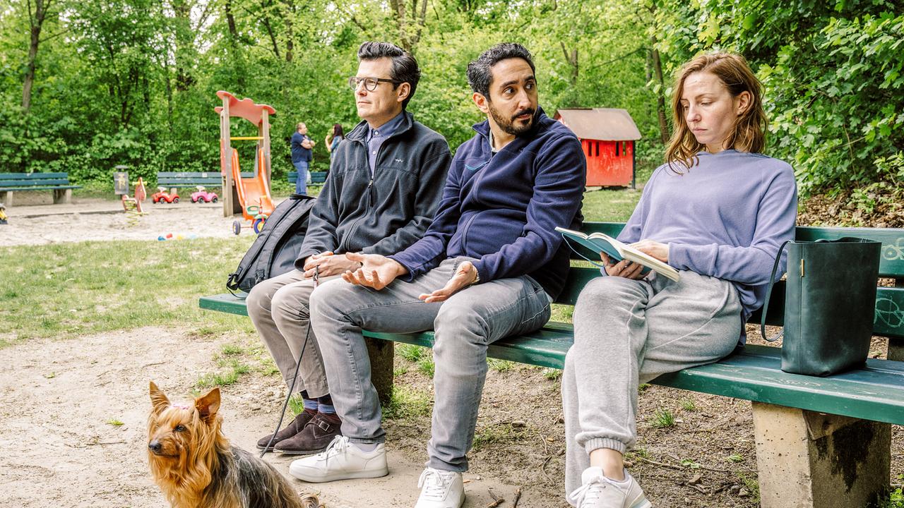 Die Glücksspieler Jasper, Firat und Ines sitzen nebeneinander auf einer Parkbank neben einem Spielplatz.