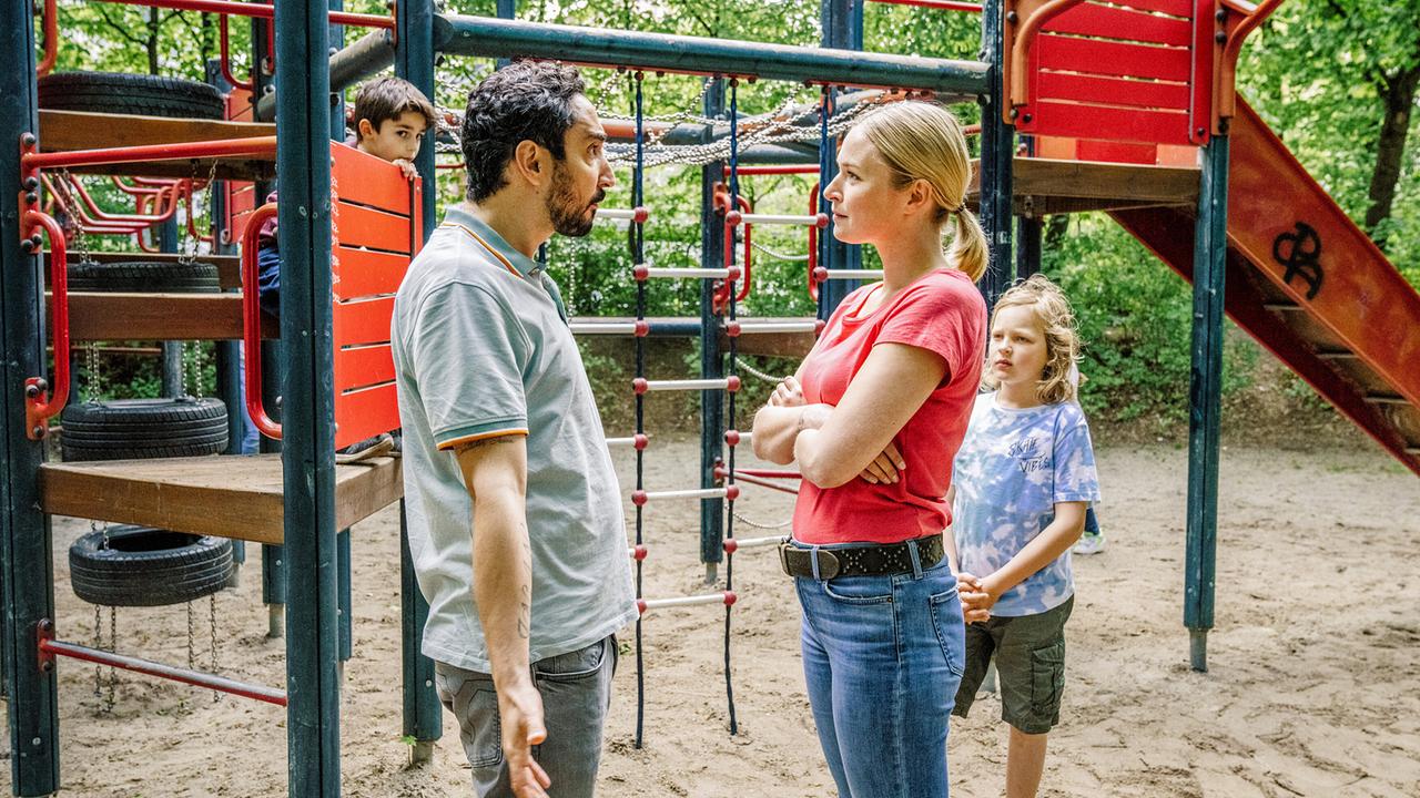 Firat streitet auf dem Spielplatz zwischen Klettergerüsten mit Tristans Mutter, weil ihr Sohn seinen Sohn Hugo nicht auf die Schaukel lässt.
