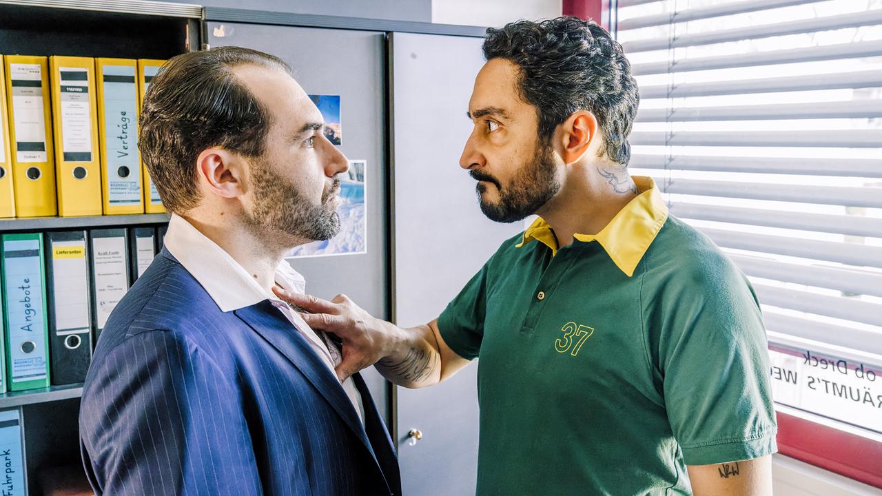 In einem engen Büro vor einem Ordnerschrank greift Firat wütend Ömer an seinem Revers.