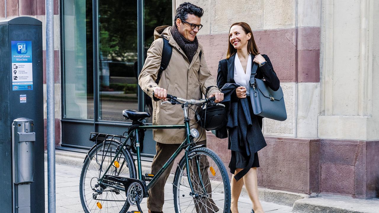 Jasper schiebt sein Fahrrad neben Ines auf einem Gehsteig.