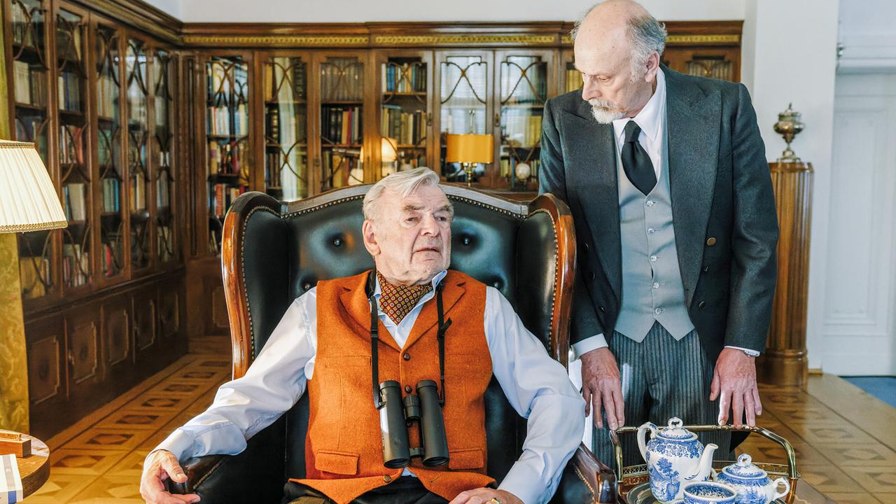 Herr Wagner unterrichtet Herrn Herzinger, der mit einem Fernglas um den Hals in einem großen Ledersessel in einem elegant eingerichteten Raum sitzt, über die neuesten Ereignisse.