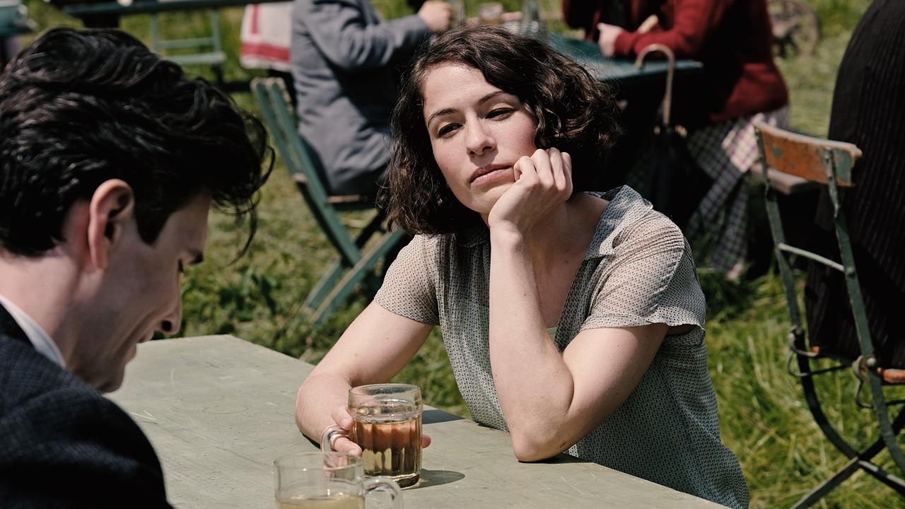 Dora sitzt nachdenklich an einem Tisch in einem Gastgarten im Freien mit einem Glas in der Hand und blickt zu Franz Kafka ihr gegenüber.