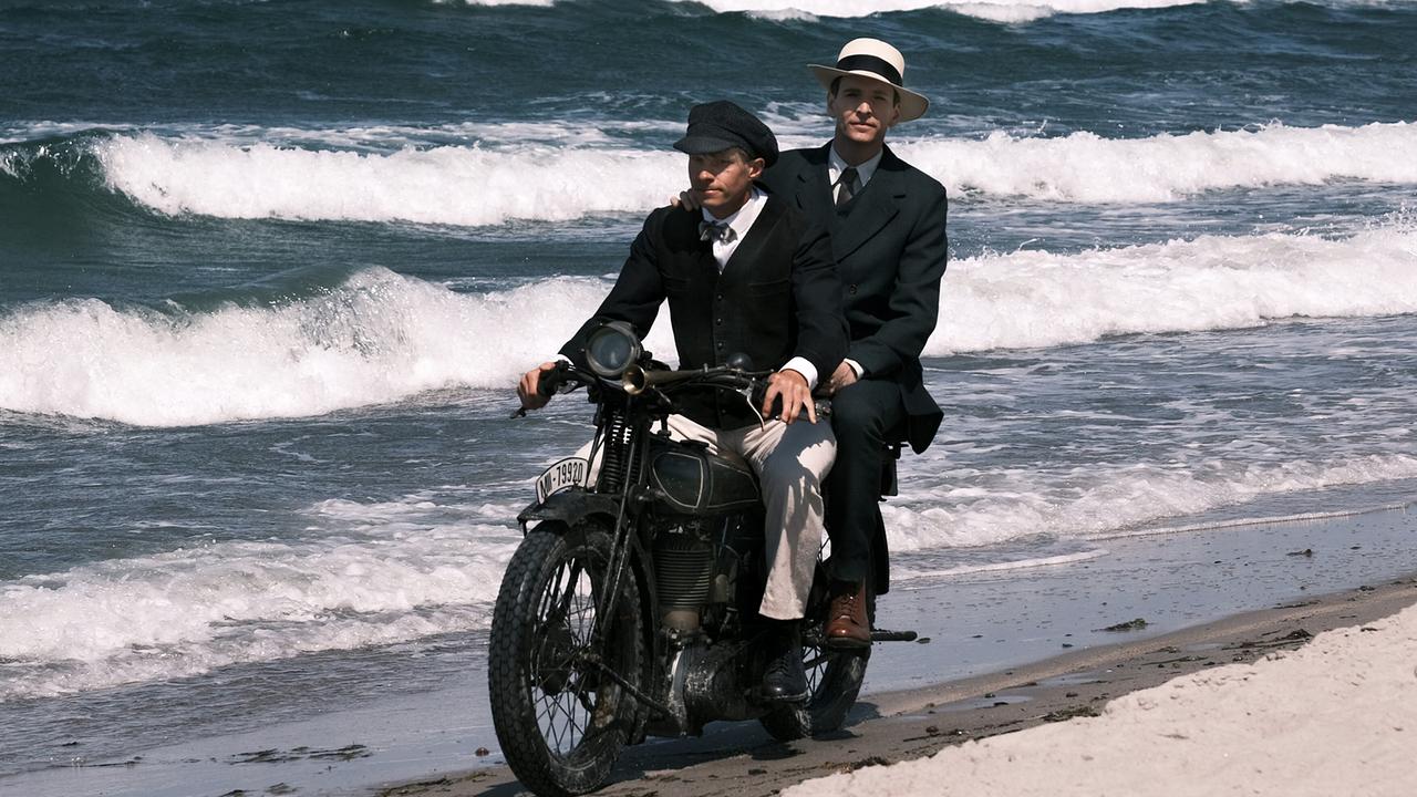 Franz Kafka am Soziussitz fährt mit einem Mann mit einem Motorrad am Strand entlang, das Meer im Hintergrund.