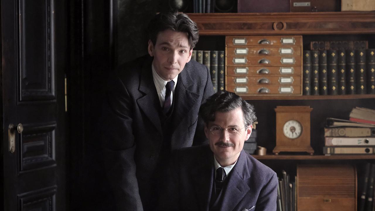 Franz Kafka und Max Brod stehen bzw. sitzen an einem Schreibtisch in einem Büro und schauen nach vorne.