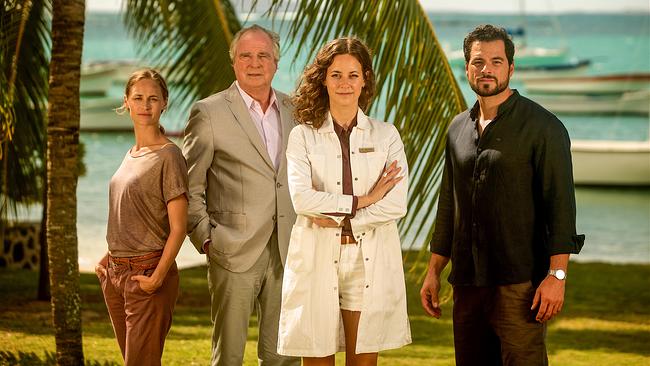 Zwei Frauen und zwei Männer stehen nebeneinander vor einer Palme, im Hintergrund sind Meer, Strand und Segelboote zu sehen