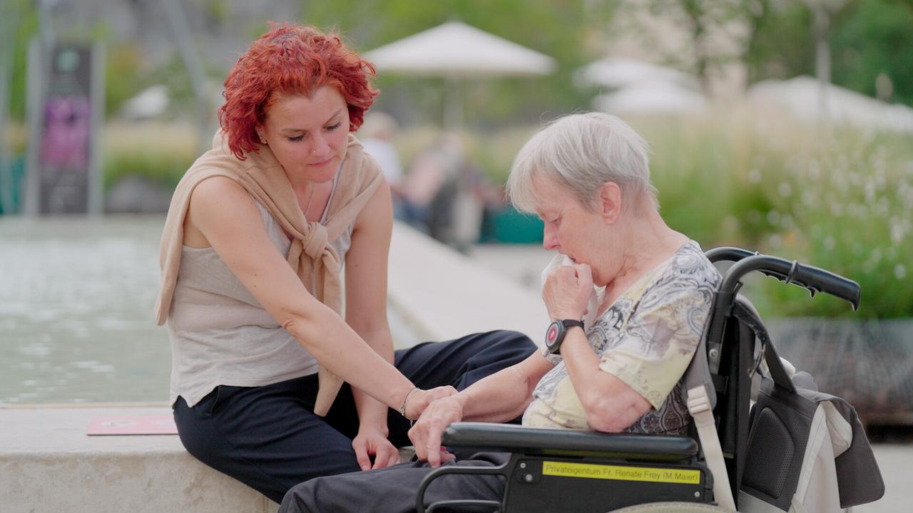 Konzertgeigerin Iris Krall-Radulian sitzend am Rand eines Wasserbeckens berührt vorlehnend den Arm von einer Caritas Socialis-Bewohnerin im Rollstuhl