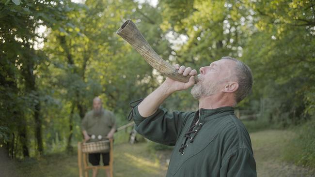 © ORF/Feature Filmproduktion/Fritz Kalteis Ein Mann bläst zur Sommersonnenwende im Grünen ein Horn, während im Hintergrund ein weiterer Mann an einer Trommel steht.
