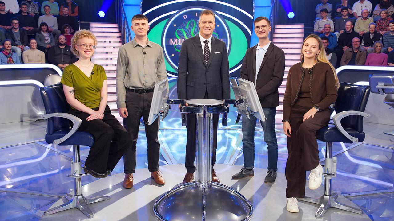 Lea Schön, Jonas Fischer, Armin Assinger, Sebastian Hopfmüller und Julia Helm stehen im "Die Millionenshow"-Studio  vor einem zentralen Moderationstisch, während Publikum auf den Rängen zusieht.