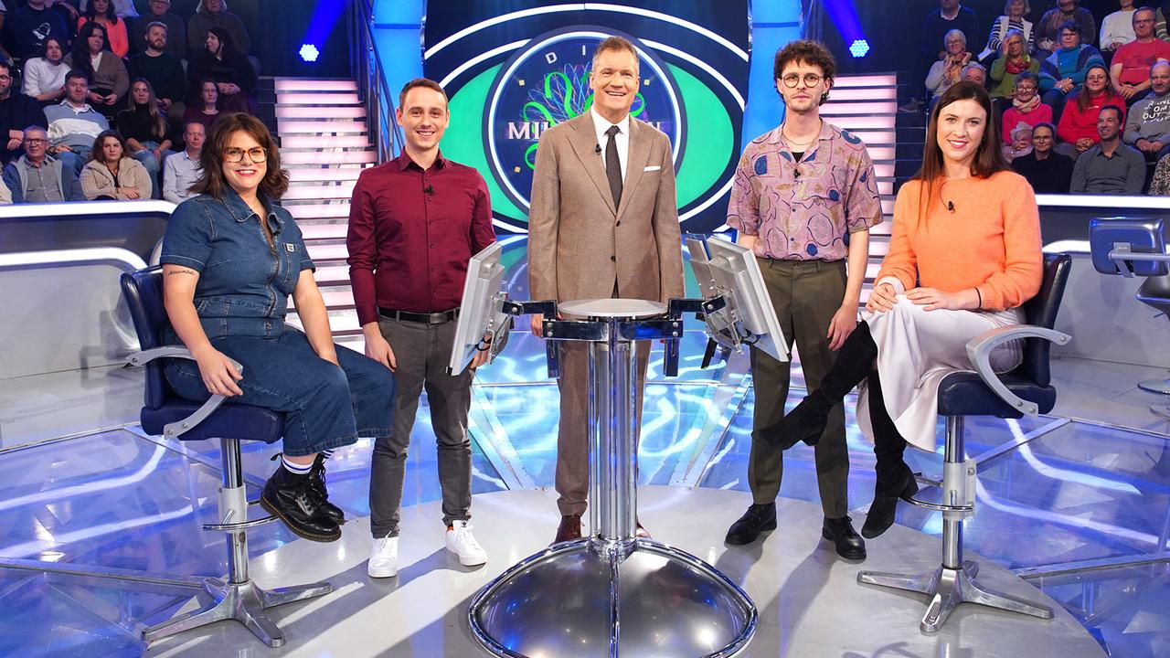 Melanie Zeiler-Rausch, Simon Pojer, Armin Assinger, Adrian Fleisch und Veronika Möller sitzen bzw. stehen um den Pult im "Die Millionenshow"-Studio mit dem  Publikum im Hintergrund.