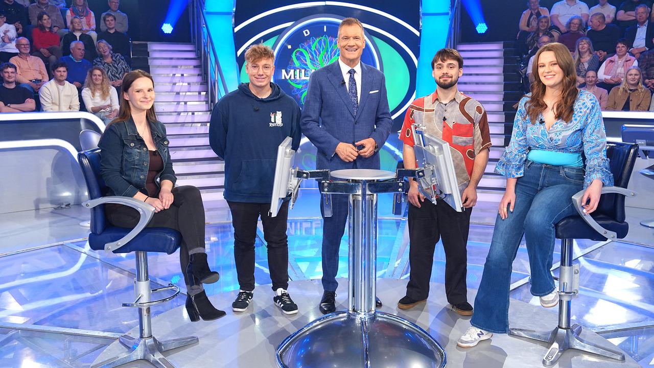 © ORF/Stefan Gregorowius Valerie Eberle, Moritz Voglmeir, Armin Assinger, Maximilian Braun und Olivia Leopold stehen gemeinsam um den "Millionenshow"-Pult im TV-Studio mit Publikum im Hintergrund.