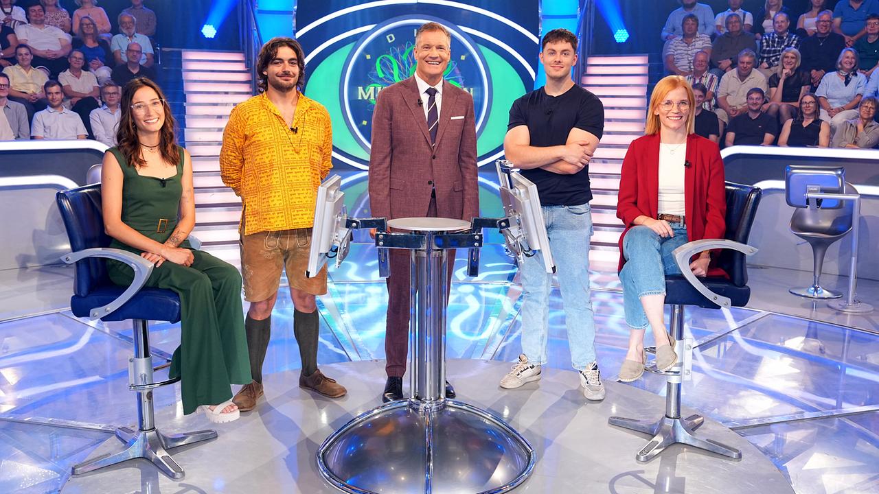 © ORF/Stefan Gregorowius Julia Avi, Manuel Rom, Armin Assinger, Philipp Bogner und Nina Schwaiger stehen lächelnd um den "Millionenshow"-Pult im TV-Studio mit Publikum im Hintergrund.