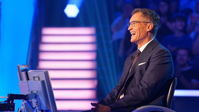Der lachender Armin Assinger im Anzug sitzt in einem blau beleuchteten " Die Millionenshow"-Studio vor einem Monitor, im Hintergrund ist Publikum zu sehen.