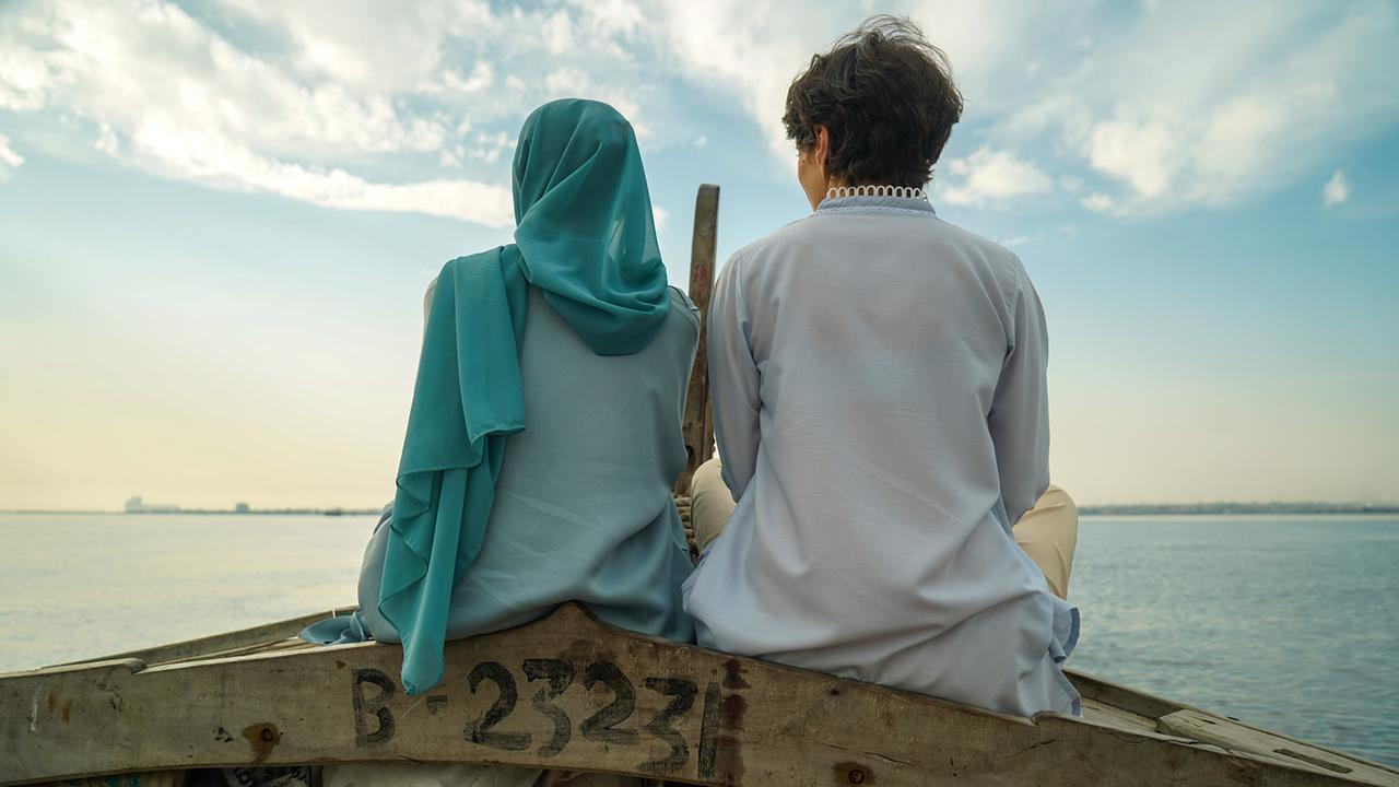 Ansicht von hinten von Rosa Lyon im Gespräch mit Muqaddas Tahir, die im Bug eines kleinen Holzbootes sitzen und über ruhiges Wasser in den wolkigen Himmel blicken.