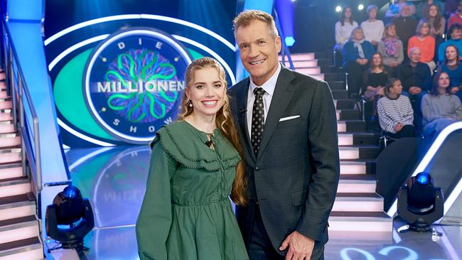 Anna Strigl und Armin Assinger posieren im Millionenshow-Studio vor dem Logo der Show, während das Publikum im Hintergrund sitzt.