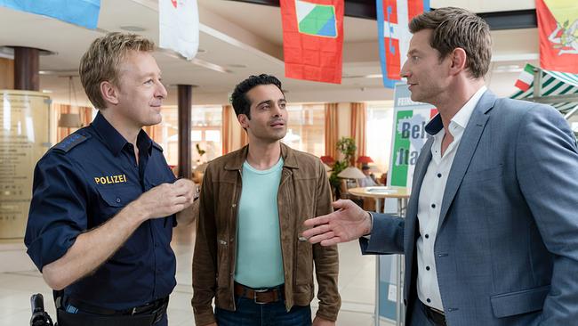 Polizeihauptmeister Mohr gibt den Kommissaren Kaya und Hansen in einer hellen Lobby bei der Feinkostmesse erste Informationen zum aktuellen Mordfall.