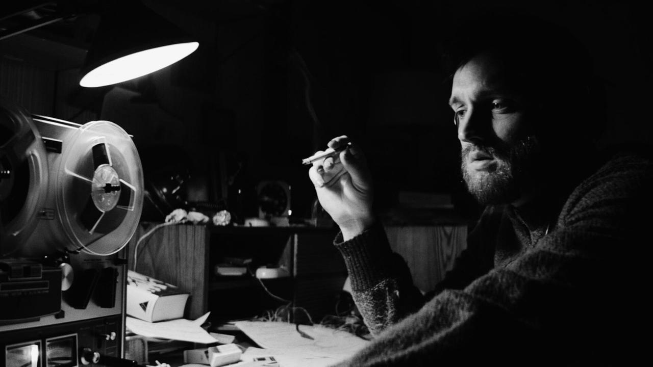 Johannes sitzt nachdenklich an einem Schreibtisch mit Tonbandgerät und hält eine Zigarette in der Hand.