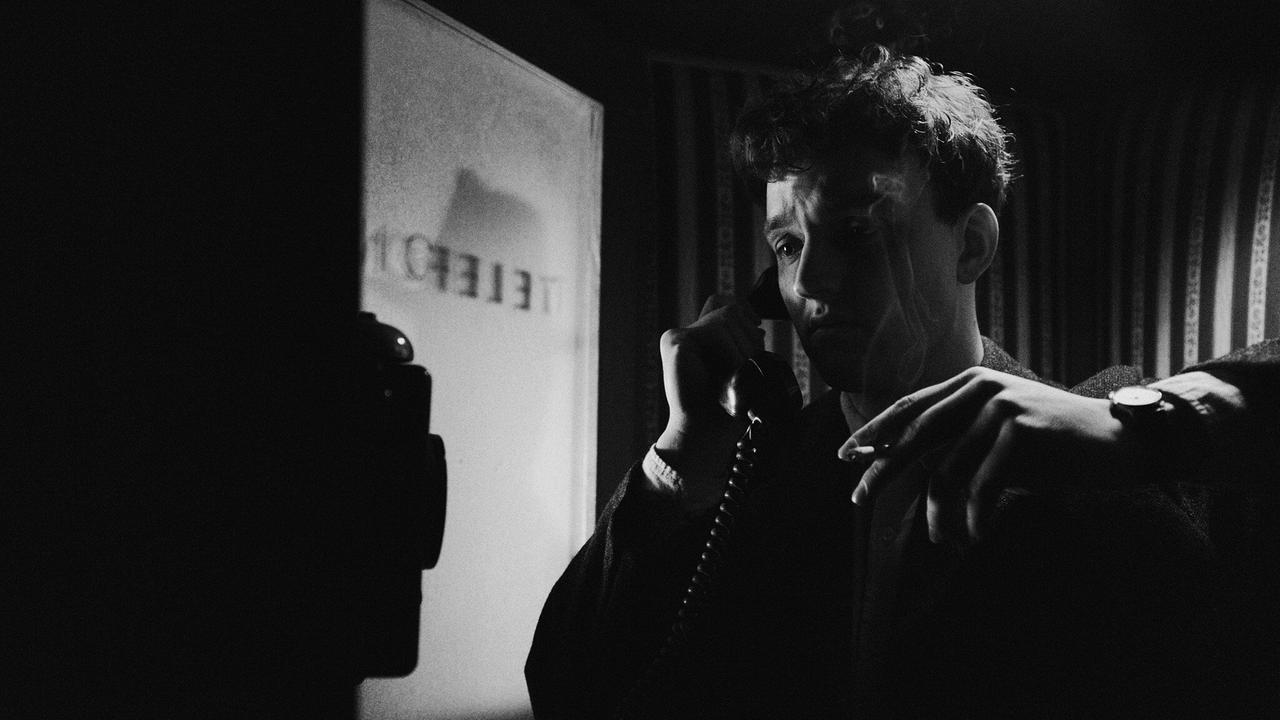 Johannes telefoniert rauchend mit seiner Mutter in einem dunklen Raum an einem Wandtelefon