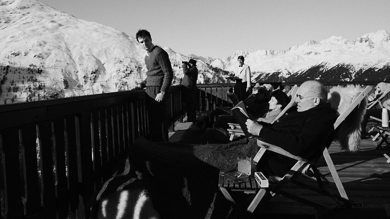 Johannes steht auf der Sonnenterasse mit Blick auf eine verschneite Berglandschaft. Im Vordergrund liest sein Doktorvater Strathen, der in einem Liegestuhl sitzt, seinen Traktat.