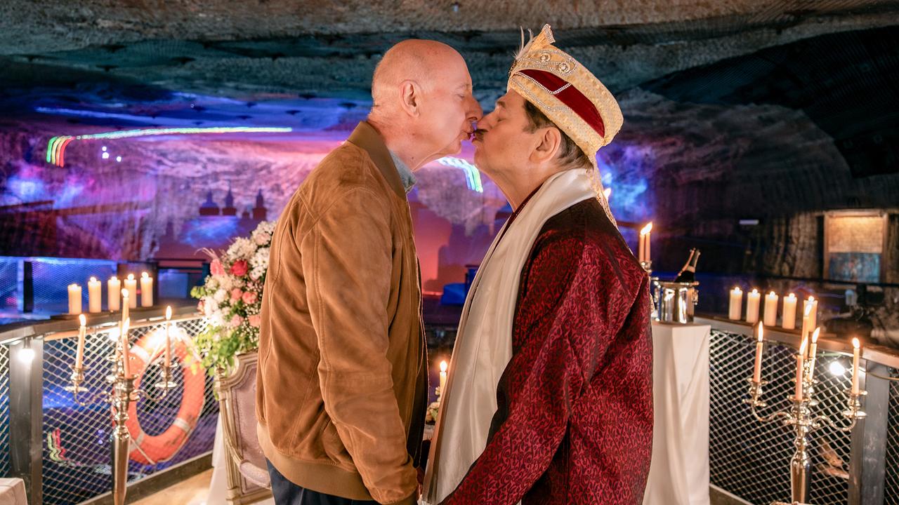 Rene Kleist und Alfons Seywald stehen sich in einem dekorierten Raum gegenüber, halten sich an den Händen und berühren ihre Lippen.