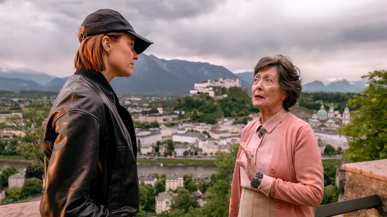 Lu und Klara Frey unterhalten sich auf einer Terrasse mit Blick auf die Stadt Salzburg und umliegende Berge.