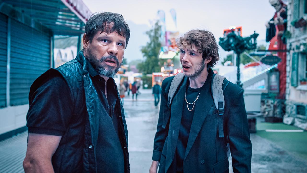 © ORF/Graf Film/Lukas Gnaiger Bruno und Tassilo Kurka stehen nebeneinander auf einem regnerischen Jahrmarkt‑Gelände