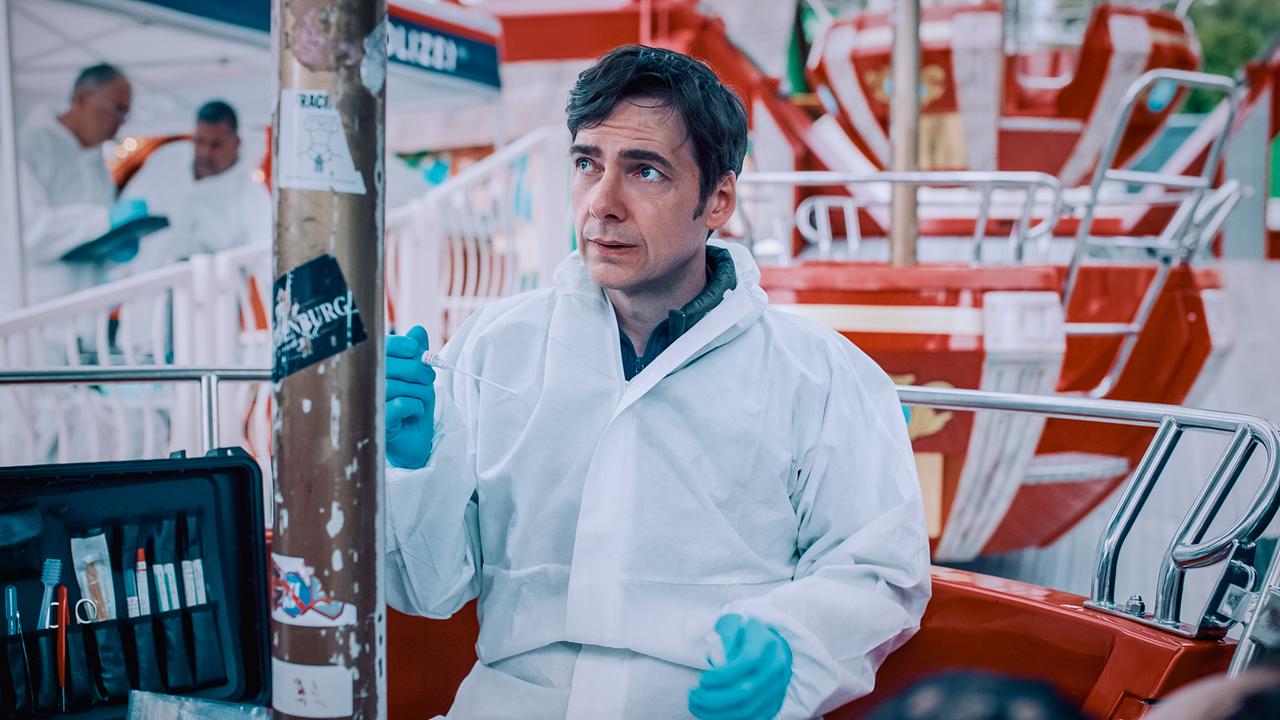 © ORF/Graf Film/Lukas Gnaiger Thomas Egger im Schutzanzug sitzt bei einer Karussell‑Bühne und führt forensische Arbeiten durch.