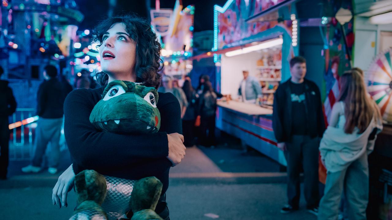 © ORF/Graf Film/Lukas Gnaiger Mara Eisler hält einen großen Plüschdrachen im Arm und blickt sich staunend auf einem farbenfrohen, nächtlichen Jahrmarkt um.