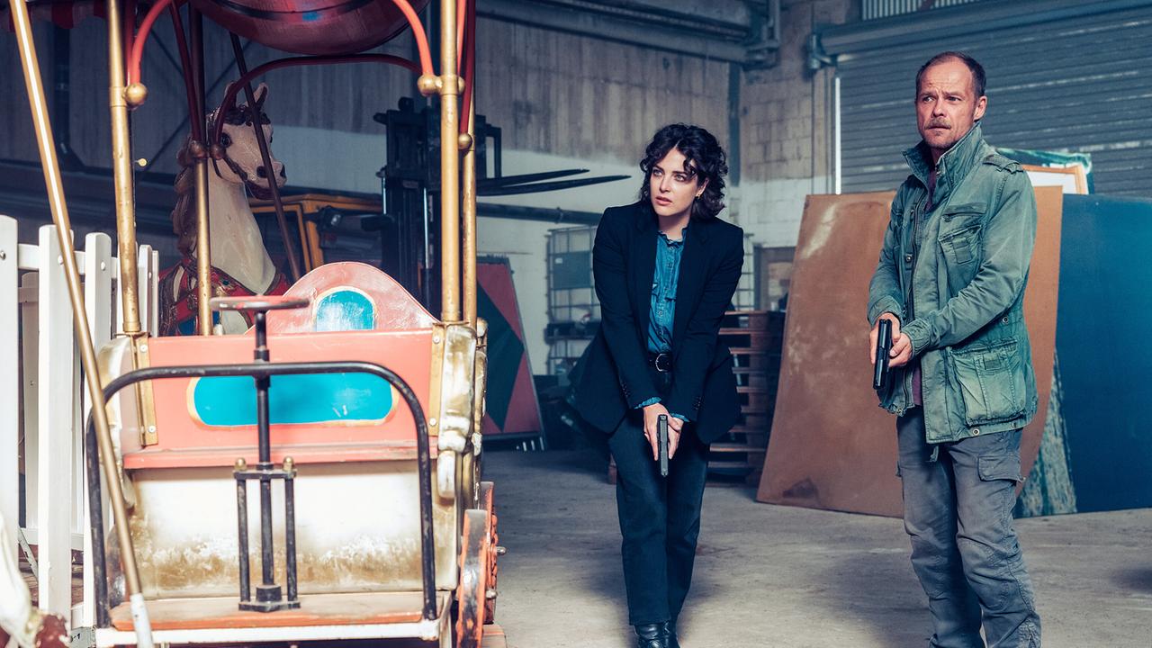 © ORF/Graf Film/Manuel Riesterer Mara Eisler und Micha Oberländer mit Pistolen in den Händen stehen in einer Lagerhalle neben einem alten Karussell.