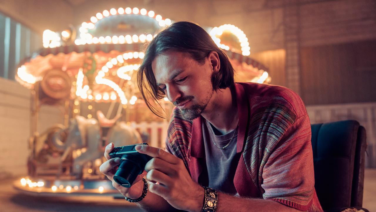 © ORF/Graf Film/Manuel Riesterer Gino Renoldi betrachtet Fotos auf einer Kamera, während hinter ihm ein beleuchtetes Karussell steht.