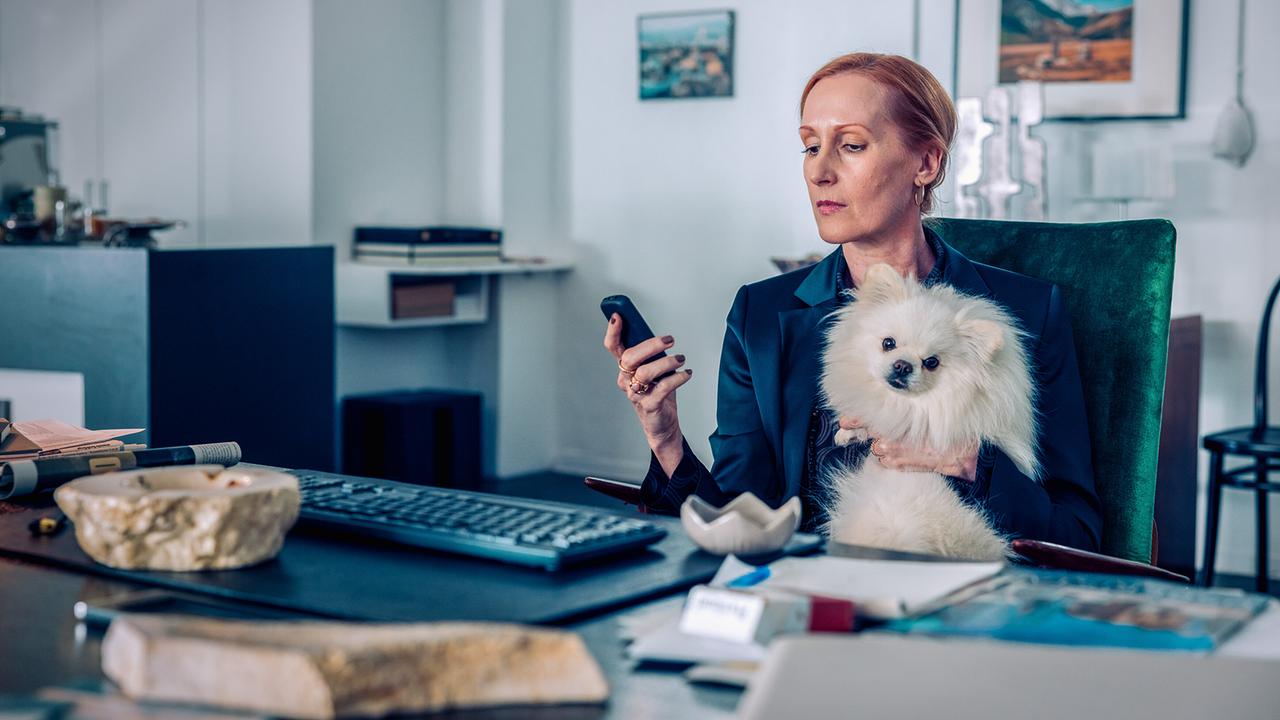 © ORF/Graf Film/Manuel Riesterer Doren Zaminsky sitzt in einem Büro an einem Schreibtisch, schaut auf ihr Handy und hält einen kleinen weißen Hund im Arm.