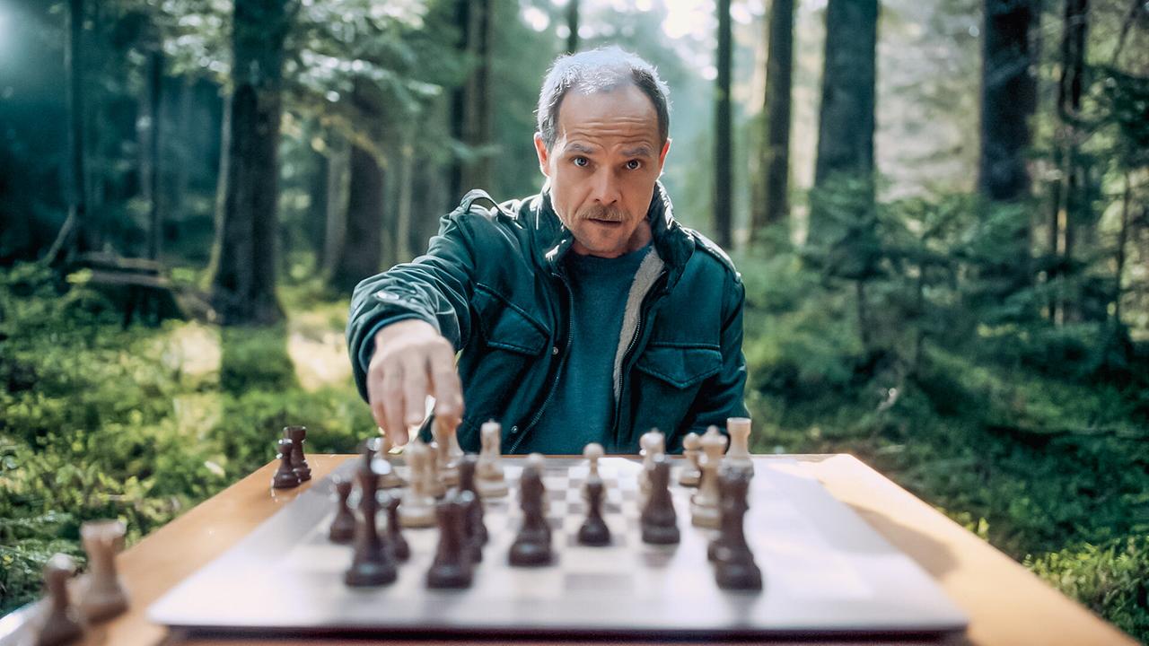 © ORF/Graf Film/Lukas Gnaiger Micha Oberländer sitzt mitten in einem sonnigen Wald an einem Tisch und spielt Schach.