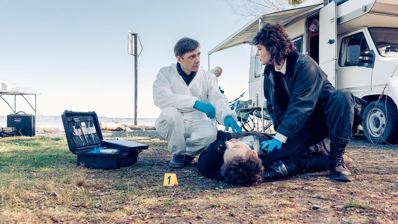© ORF/Graf Film/Manuel Riesterer Thomas Egger im Schutzanzug und Ermittlerin Mara Eisler untersuchen neben einem Wohnmobil den leblosen Andreas Janko, während eine Spurensicherungstafel im Gras steht.