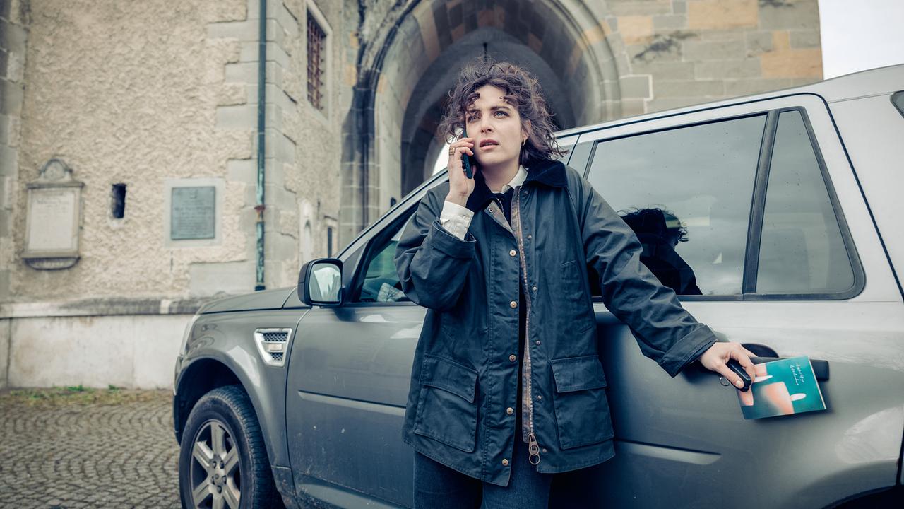 © ORF/Graf Film/Manuel Riesterer Mara Eisler telefoniert nervös neben einem Wagen vor einem Kirchenportal.