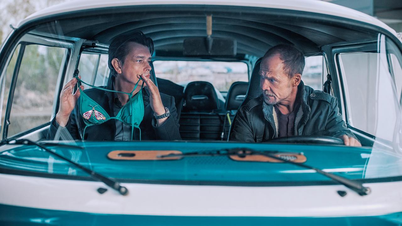 © ORF/Graf Film/Lukas Gnaiger Thomas Komlatschek sitzt neben Micha Oberländer in einem Kleinbus und hält ein türkisfarbenes Bikinioberteil in der Hand.