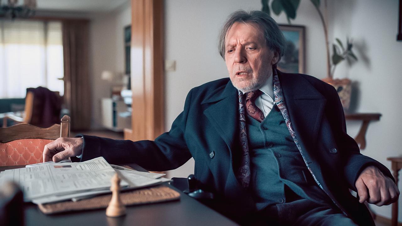© ORF/Graf Film/Lukas Gnaiger Joseph Hadeling sitzt an einem Tisch mit Unterlagen und schaut auf eine Schachfigur vor ihm auf dem Tisch.
