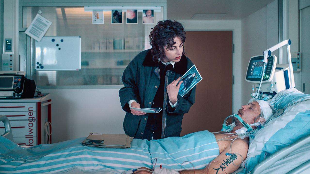© ORF/Graf Film/Lukas Gnaiger Ermittlerin Mara Eisler steht am Krankenbett von Micha Oberländer und begutachtet Fotos.